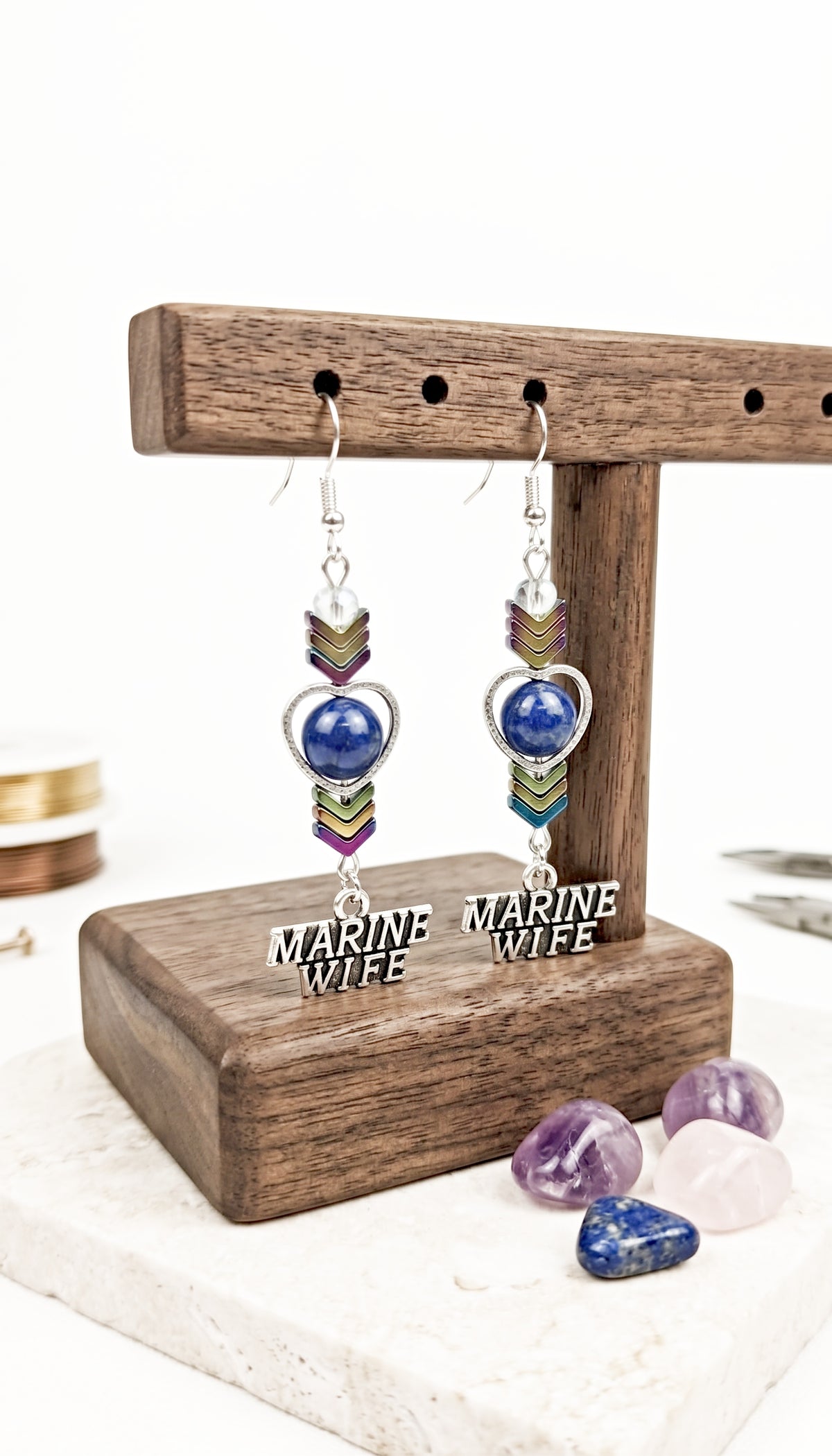A pair of dangling earrings displayed on a wooden stand. Each earring has a silver fishhook, a silver heart outline, a blue oval stone, colorful chevron beads in green, purple, and gold, and a silver dangling tag that reads "MARINE WIFE" in silver letters.
