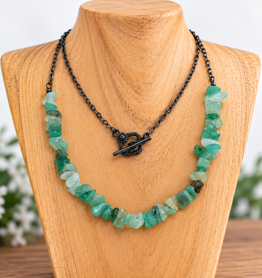 A chunky gemstone necklace made of green translucent stones on a black chain with a toggle clasp, displayed on a wooden jewelry bust.