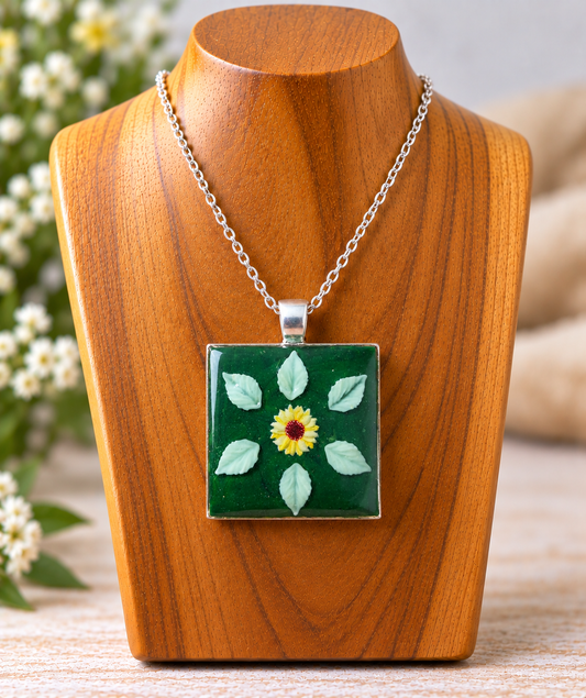 A silver chain necklace with a square pendant featuring a green background, a yellow flower in the center, and six light green leaves, displayed on a wooden bust.