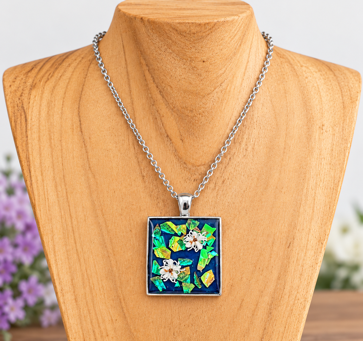 A square pendant necklace displayed on a wooden mannequin bust. The pendant features a colorful mosaic design with blue, green, yellow, and white floral elements, suspended from a silver chain.