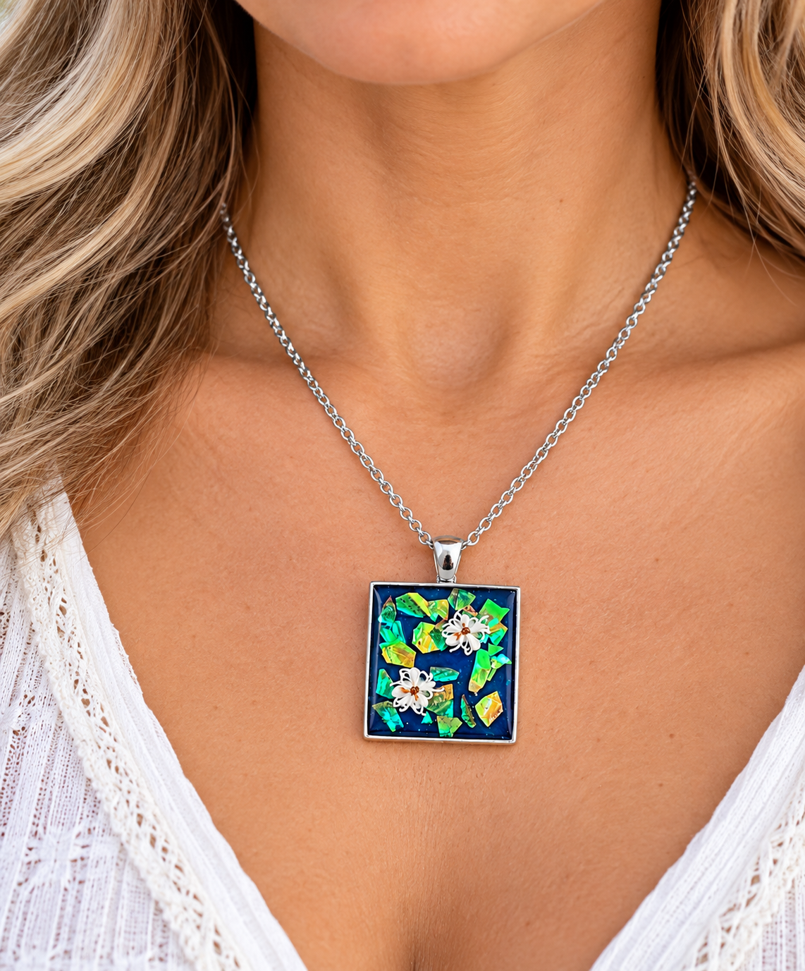 A close-up of a model wearing a silver chain necklace with a square pendant featuring a colorful floral design on a dark blue background.
