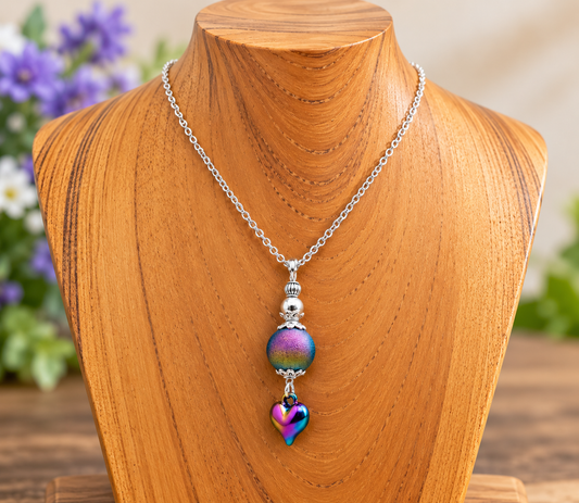 A silver chain necklace with a round iridescent bead and a dangling heart charm, displayed on a wooden jewelry bust.