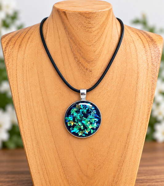 A round pendant necklace with a black cord and a silver-toned bezel, featuring iridescent blue, green, and gold leaf-like flakes on a dark background, displayed on a wooden mannequin bust.