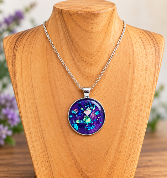 A silver chain necklace with a round pendant featuring a purple and blue iridescent mosaic design, displayed on a wooden jewelry bust.