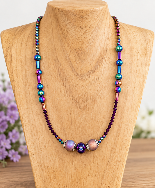 A beaded necklace displayed on a wooden jewelry bust. The necklace features iridescent metallic beads in blue, green, purple, and gold, accented by dark purple faceted beads and three larger iridescent lavender beads at the center with a small blue evil eye charm.
