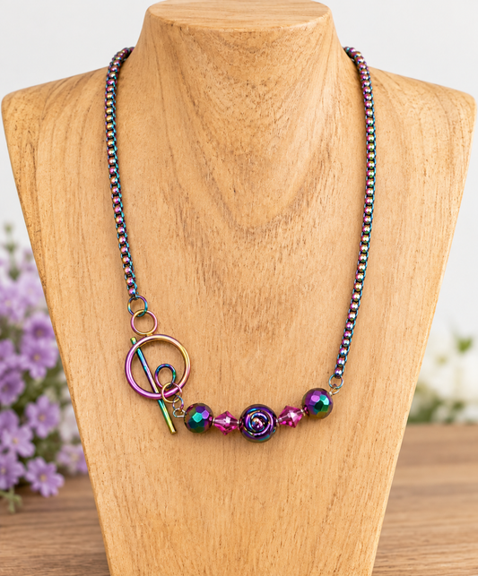A colorful beaded necklace displayed on a wooden bust. The necklace features iridescent metallic beads, a central purple rose charm, a purple bead, and a silver-tone toggle clasp with a peace sign.