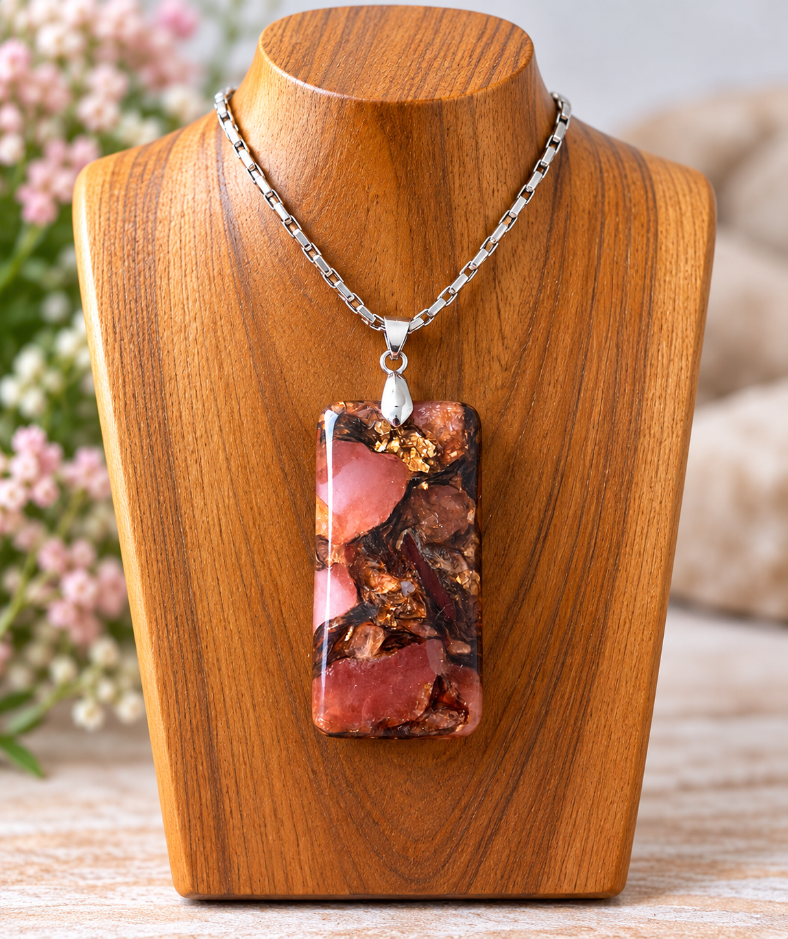 A silver chain necklace with a rectangular pendant featuring a marbled pattern of pink, brown, and gold flecks, displayed on a wooden bust.