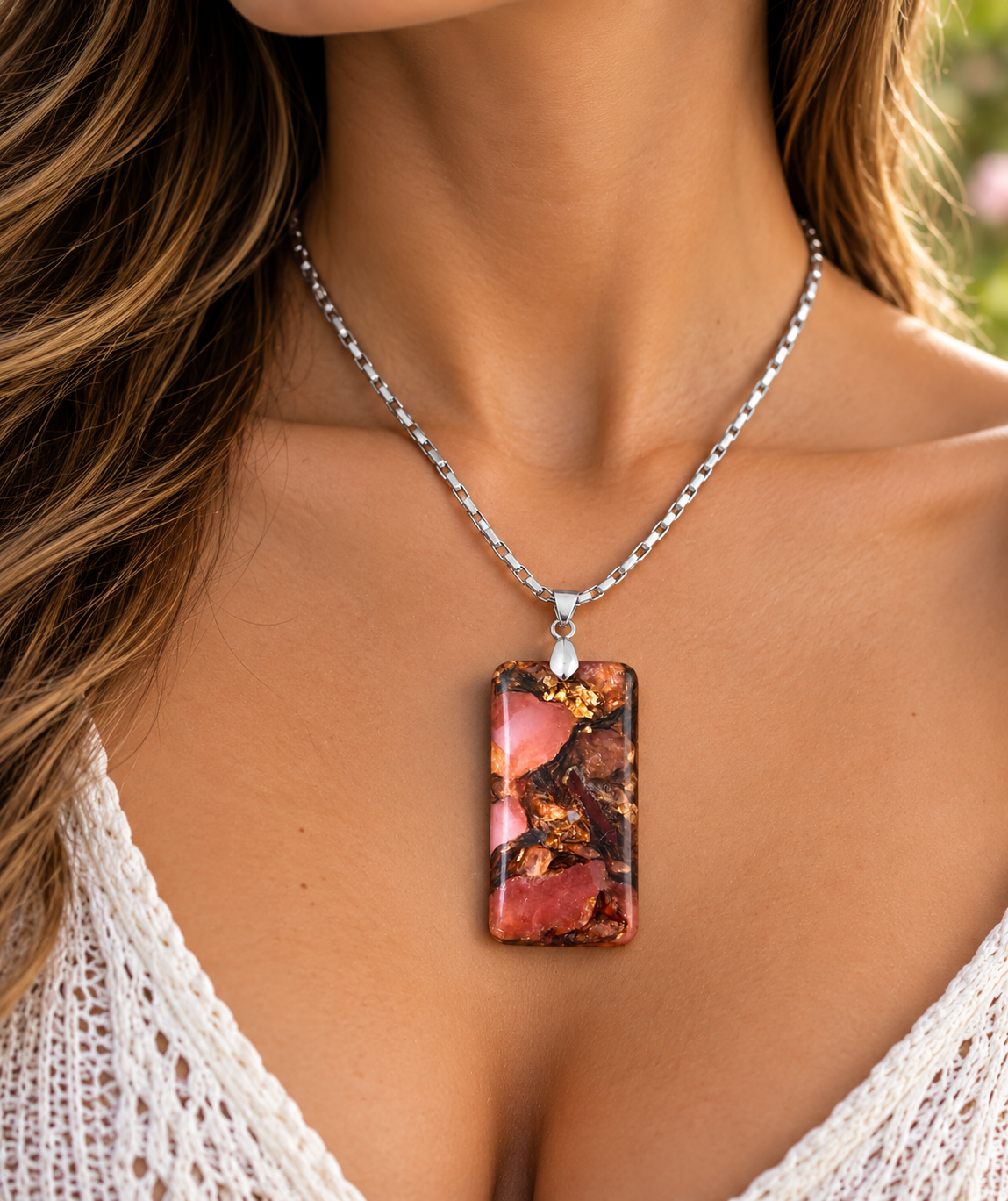 A close-up of a woman wearing a silver chain necklace with a rectangular pendant featuring a pink, red, and gold marbled pattern.