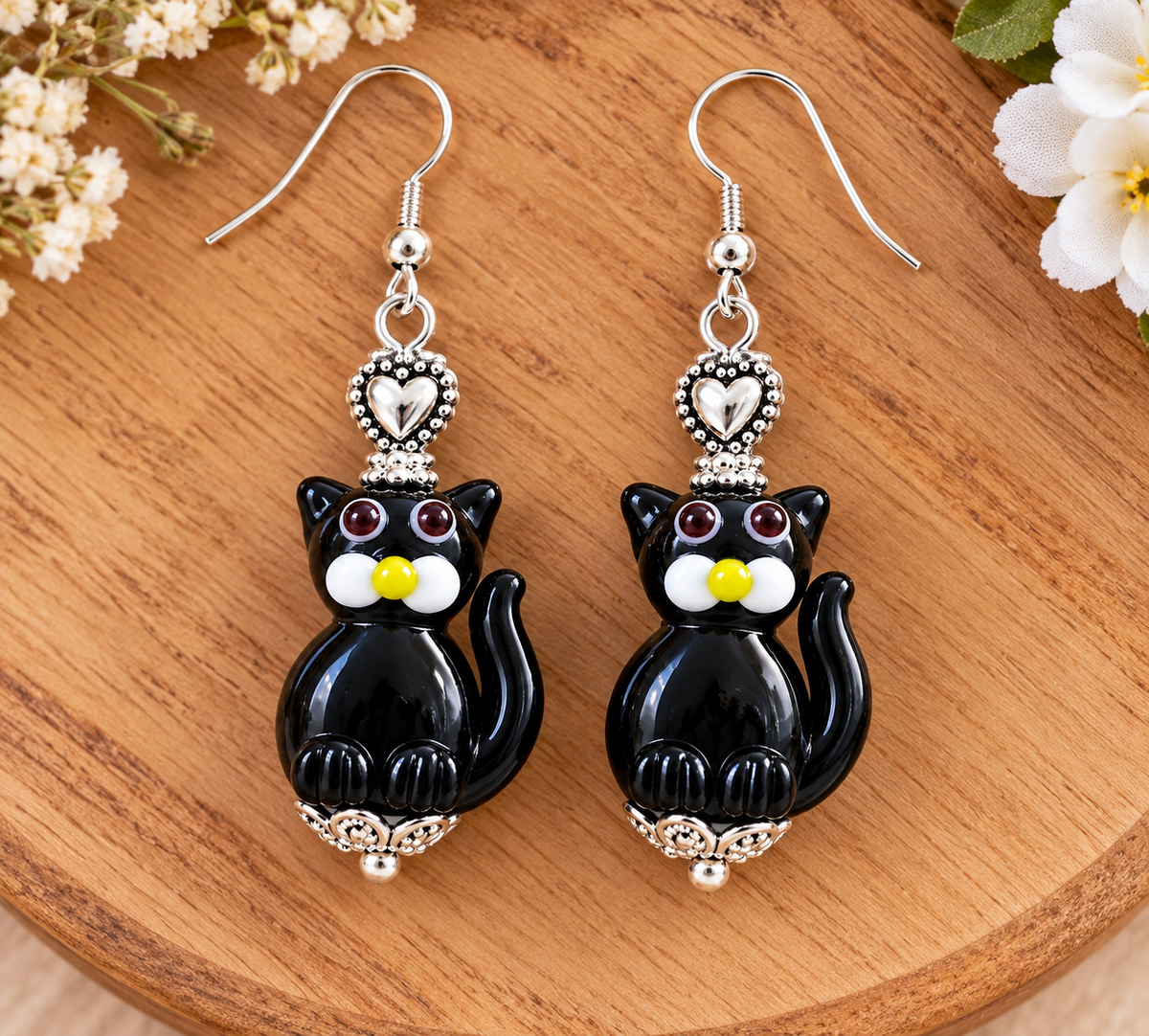 A pair of dangling cat-shaped earrings with glossy black bodies, silver-tone hooks and trim, white facial details, and small yellow accents, displayed on a wooden surface with flowers in the corners.
