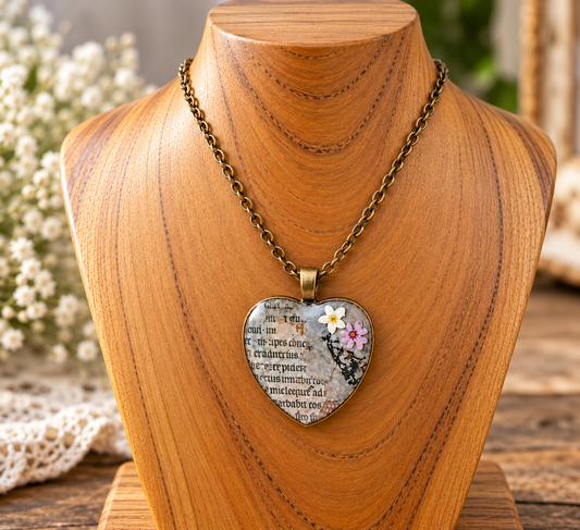 Heart-shaped pendant necklace displayed on a wooden bust. The pendant features a vintage book page background with a small white flower and pink flower decoration in the upper right corner, hanging from an antique bronze chain.