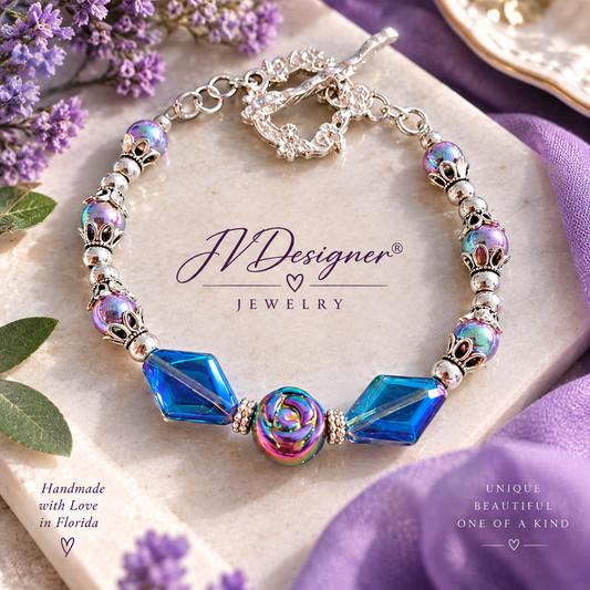 **Alt text:** `Handmade JVDesigner® jewelry bracelet with iridescent purple beads, blue faceted crystal beads, silver floral accents and rainbow rose centerpiece, displayed on a cream stone surface.`
