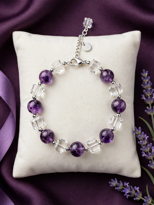 A beaded bracelet with alternating purple amethyst round beads and clear faceted crystal cube beads, displayed on a cream pillow over a purple satin background with lavender sprigs.