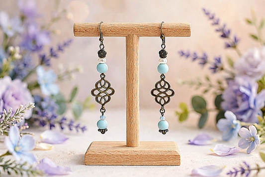A pair of dangling earrings displayed on a small wooden stand, with blurred purple and blue flowers around them. The earrings have dark metal filigree charms, light blue beads, and small clear beads.