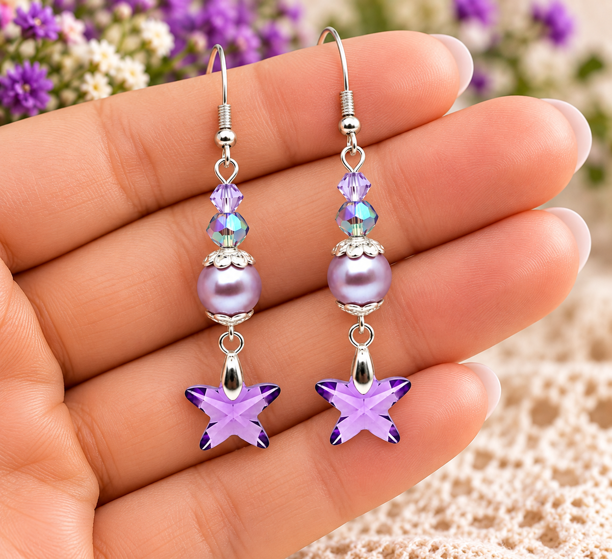 A pair of dangling earrings held in a hand. Each earring has a silver fishhook wire, a small heart-shaped crystal, two iridescent round beads, a larger lavender pearl-like bead, a silver filigree cap, and a dangling lavender crystal star charm.