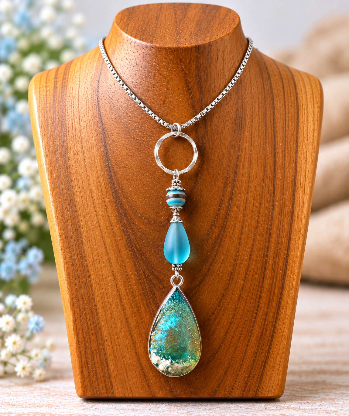 Handmade ocean resin teardrop pendant necklace with blue sea glass drop bead on titanium silver chain, displayed on a wooden bust