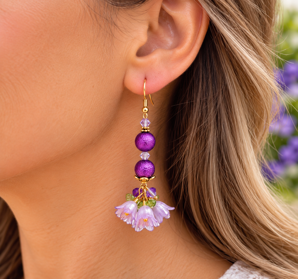 A close-up of a woman's ear wearing a dangling earring with purple beads, a gold hook, and a cluster of pale lavender bell-shaped flowers with green leaves.