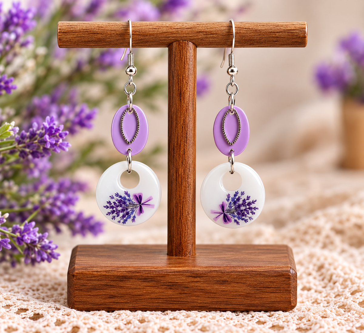 A pair of dangling earrings displayed on a wooden stand. Each earring has a lavender-purple oval top, a silver-tone fishhook ear wire, a small silver bead, and a white circular drop with a pressed purple lavender floral design.