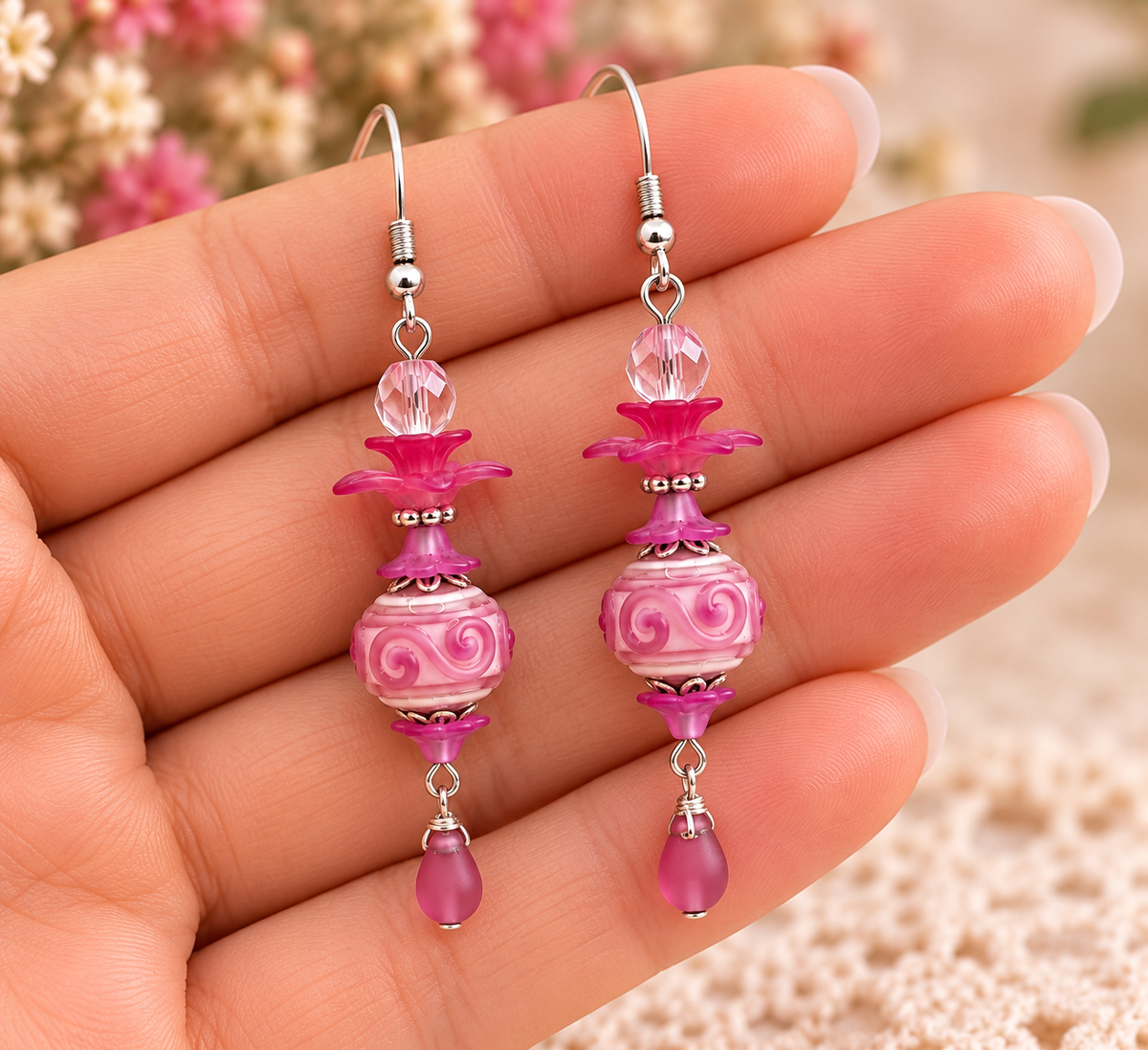 Pink and silver drop earrings held in a hand, featuring carved glass beads, floral details, and dangling teardrop charms.