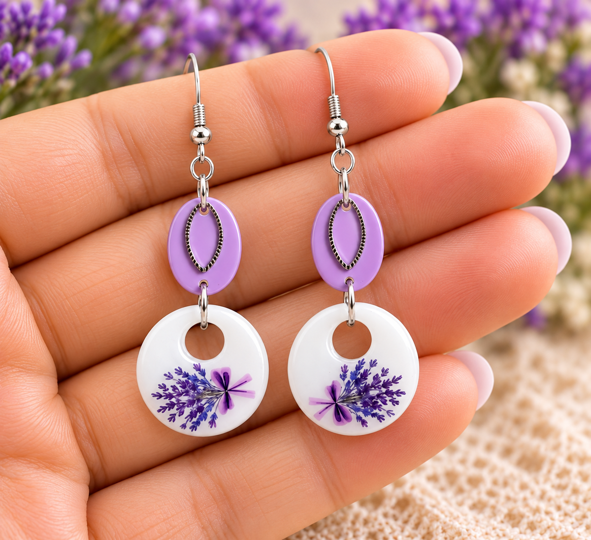 A pair of dangling earrings held in a hand, featuring silver fishhook wires, lavender-shaped oval links, and white circular drops with pressed purple lavender flowers.