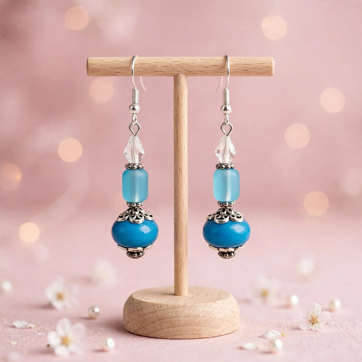 A pair of dangling earrings displayed on a small wooden stand against a soft pink bokeh background. Each earring has a silver fishhook, a clear faceted crystal, a light blue rectangular bead, a turquoise blue round bead, and an ornate silver filigree cap with a larger round turquoise blue bead below.