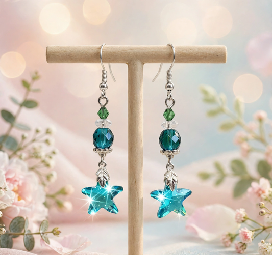 A pair of dangling earrings displayed on a small wooden stand against a soft pastel floral background. Each earring has a silver fishhook, a small green faceted bead, a larger teal-blue faceted bead, a silver leaf-shaped spacer, and a star-shaped turquoise-blue crystal at the bottom.