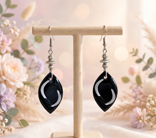 A pair of black teardrop-shaped drop earrings with silver-toned hooks and spacer beads, displayed on a light wooden stand against a soft, blurred floral background.