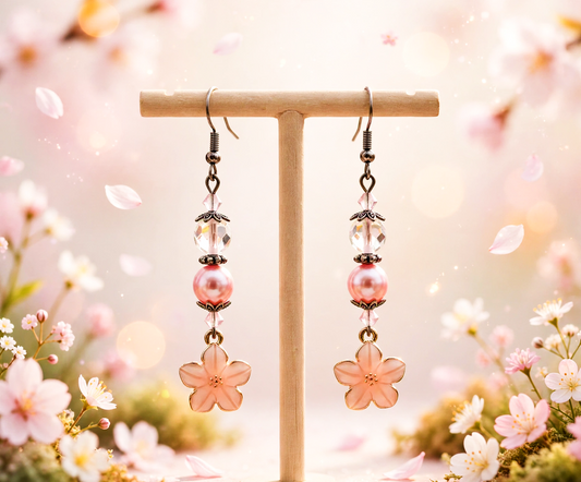 A pair of dangling earrings displayed on a small wooden T-stand. Each earring has a dark metal fishhook, a faceted clear bead, a pink pearl-like bead, a small clear bead, and a pink translucent flower charm at the bottom.