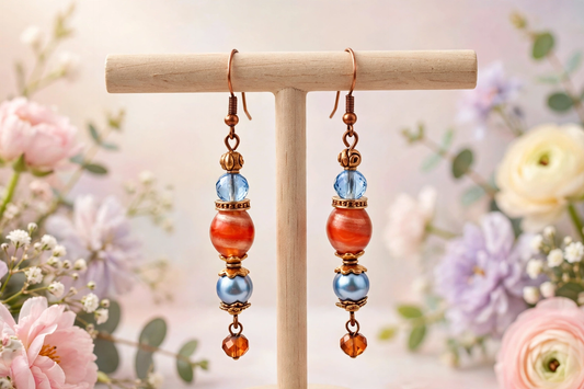A pair of handmade dangle earrings displayed on a wooden T-stand. The earrings feature copper-toned metal hooks and beads, including a light blue faceted bead, a larger orange-red marbled bead, a small gold spacer, and a light blue round bead, finished with a dangling copper bead.