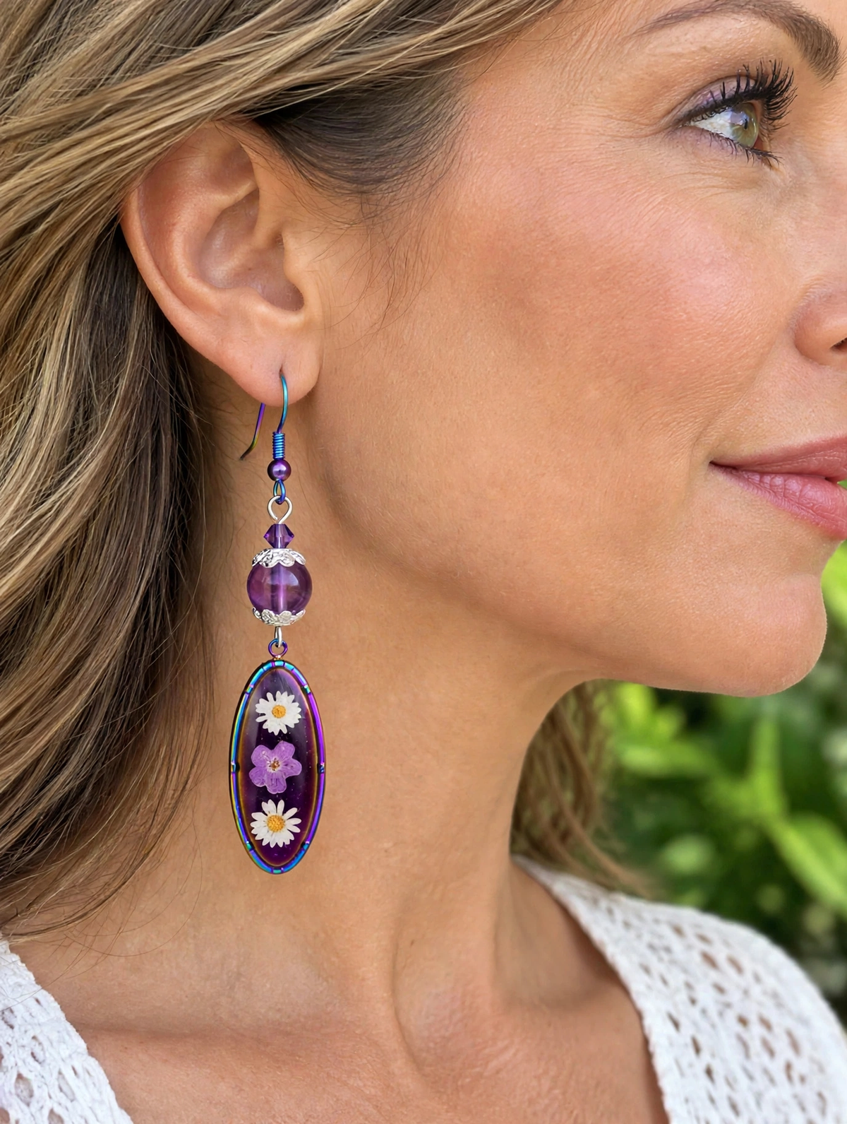 A close-up side profile of a woman wearing a dangling earring. The earring features a purple faceted bead, a silver-toned spacer, and an oval charm filled with purple and white dried flowers on a dark background.