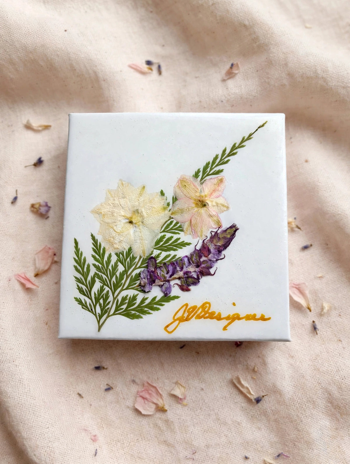 A square white ceramic tile or coaster featuring a pressed floral design with green leaves, yellow and pale pink blossoms, and purple flowers, signed in gold cursive at the bottom. It is photographed from above on a beige fabric background with scattered dried flower petals.