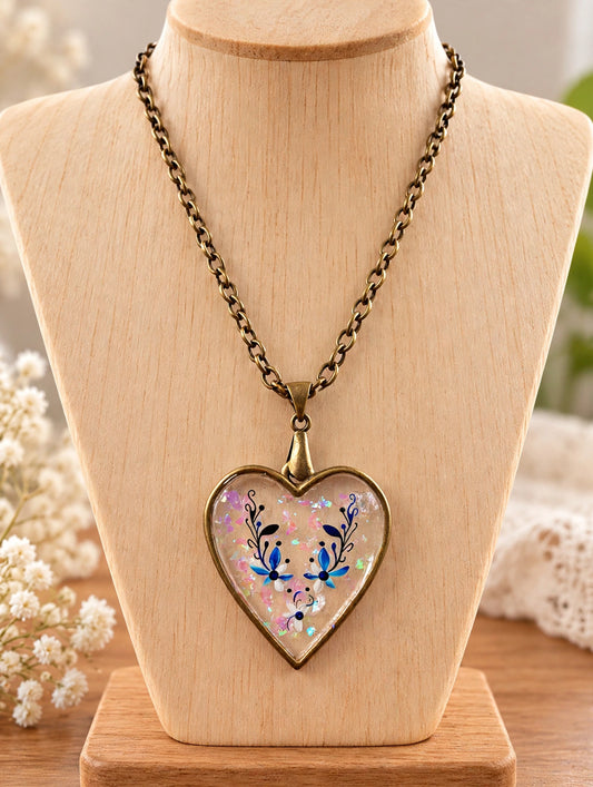 A heart-shaped pendant necklace displayed on a light wood bust. The pendant has an antique bronze border and chain, with a clear top holding iridescent glitter and blue floral details.
