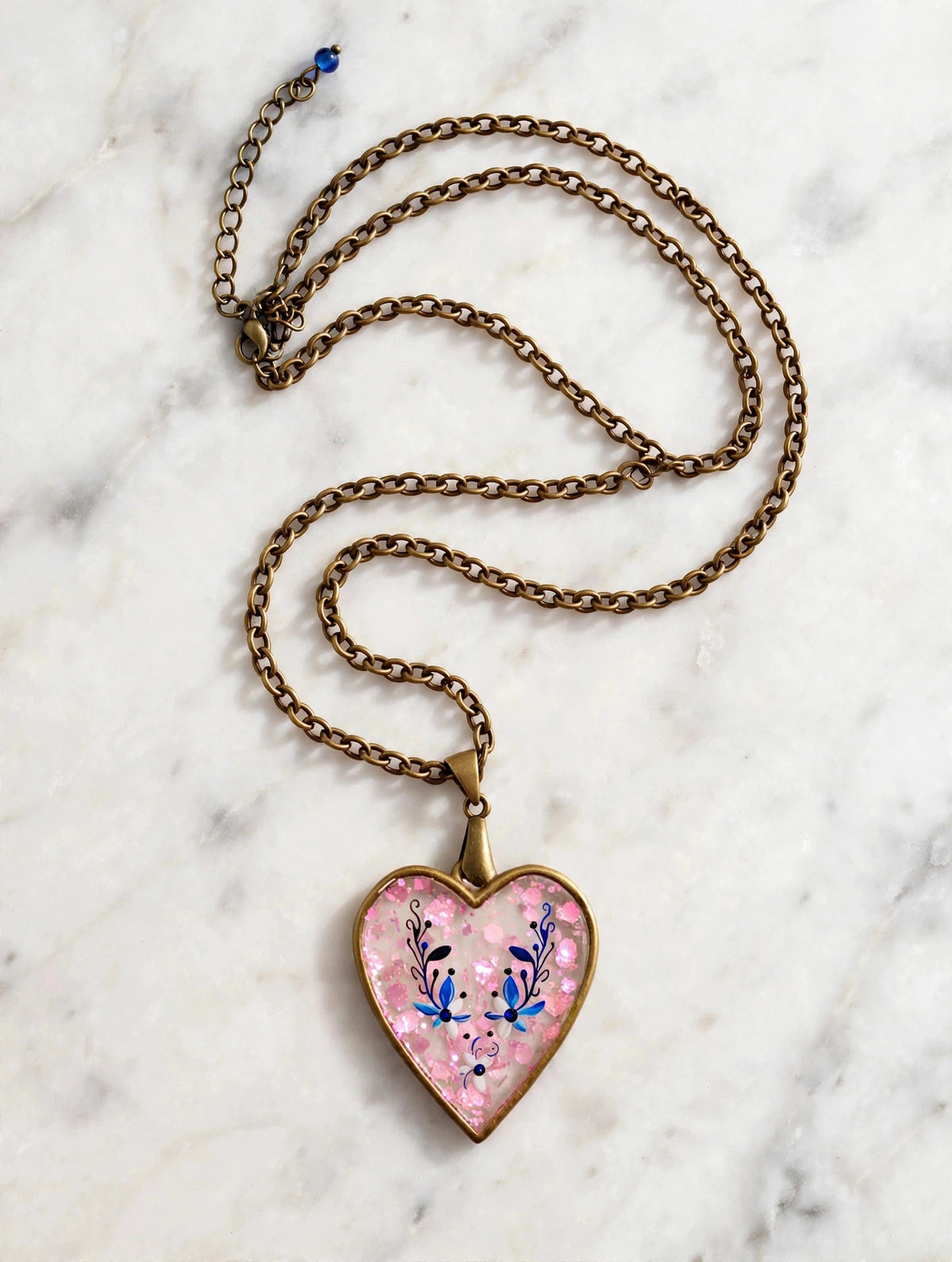A heart-shaped pendant necklace with a pink glittery background, blue floral details, and an antique bronze chain, displayed on a white marble surface.
