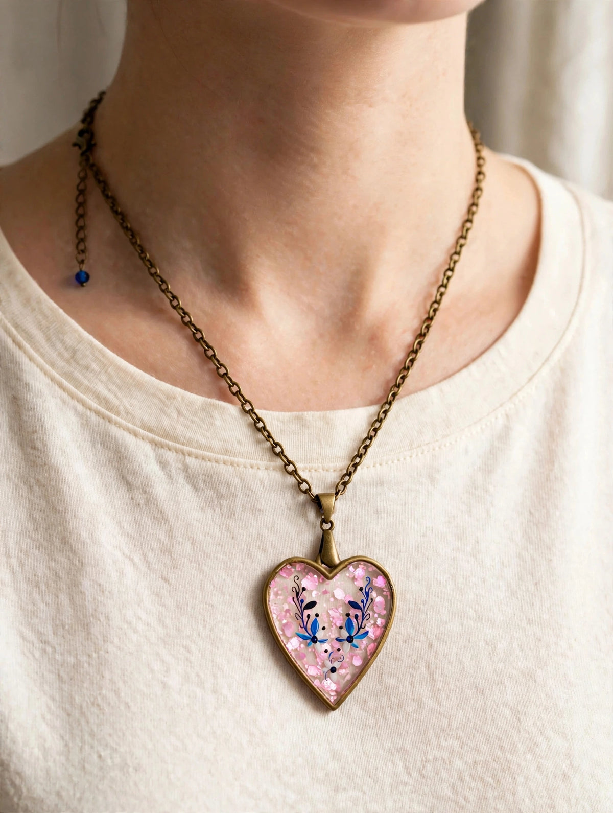 A close-up of a person wearing a heart-shaped pendant necklace. The pendant has a pink background with glitter and blue floral details, hanging from an antique bronze chain.
