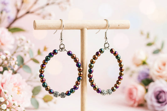 Iridescent beaded hoop earrings with silver-toned hooks and decorative silver beads, displayed on a wooden stand against a soft pink floral background.