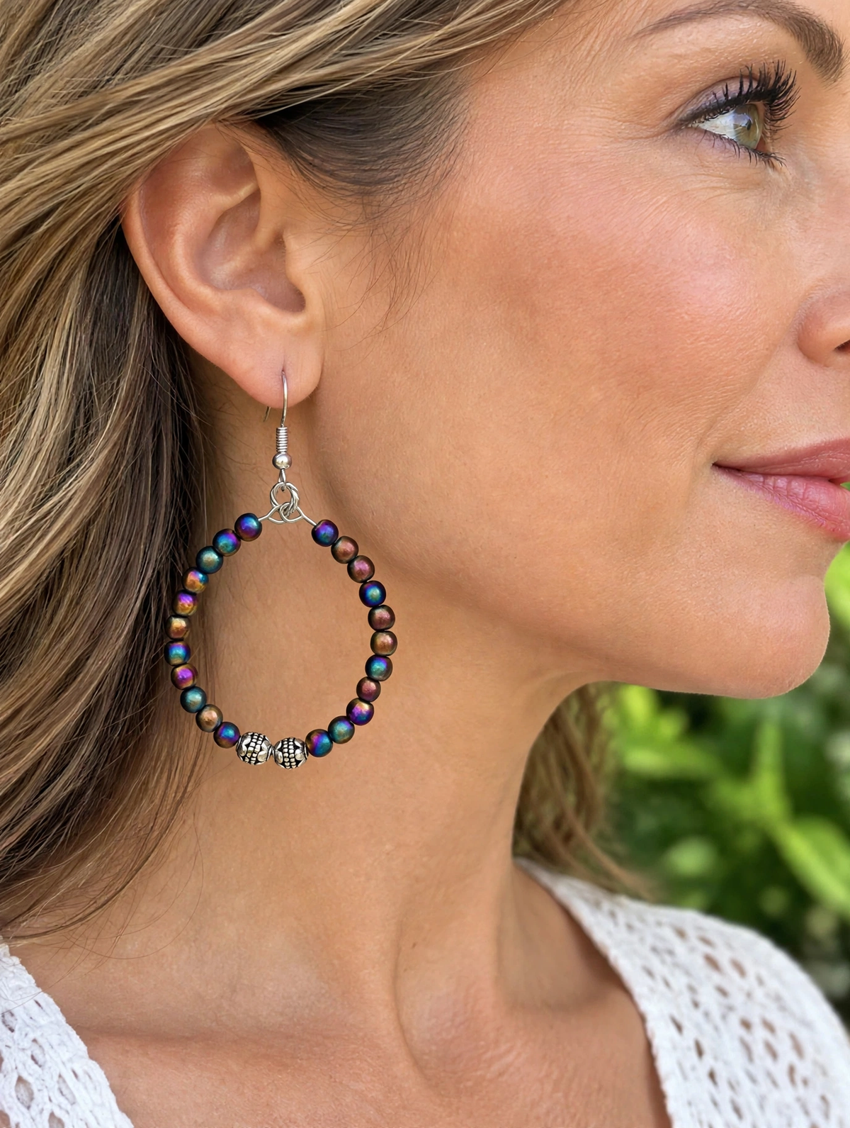 A close-up side profile of a woman wearing a large hoop earring made of iridescent dark beads with small silver charms, including a skull detail.