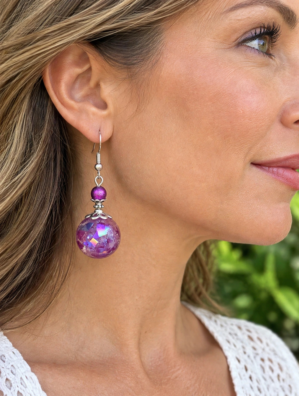 A close-up side profile of a woman wearing a dangling earring. The earring features a silver-toned fishhook, a small round purple bead, and a larger faceted purple glass bead with an iridescent finish.