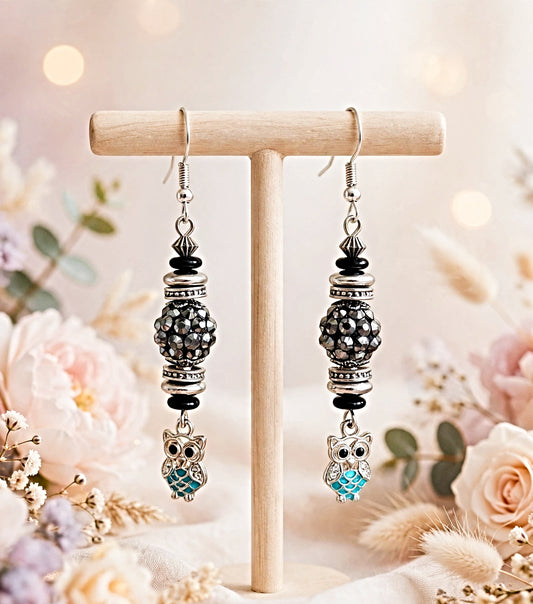 A pair of dangling owl earrings displayed on a wooden T-stand. The earrings feature silver-toned metal hooks, black spacer beads, textured black and silver bead clusters, and dangling owl charms with one light blue owl and one silver owl with black details.