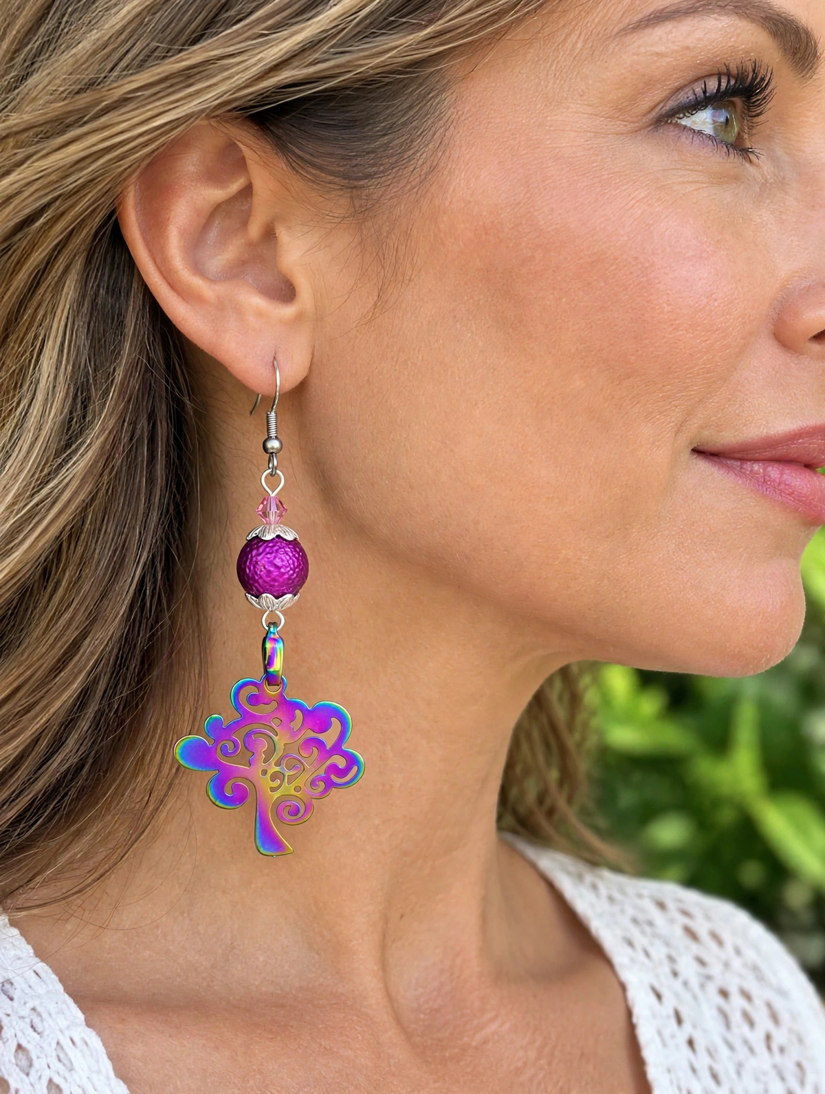 A close-up side profile of a woman wearing a dangling earring. The earring has a silver fishhook wire, a textured magenta-purple bead, and a dangling iridescent floral charm that shifts between blue, green, and purple.