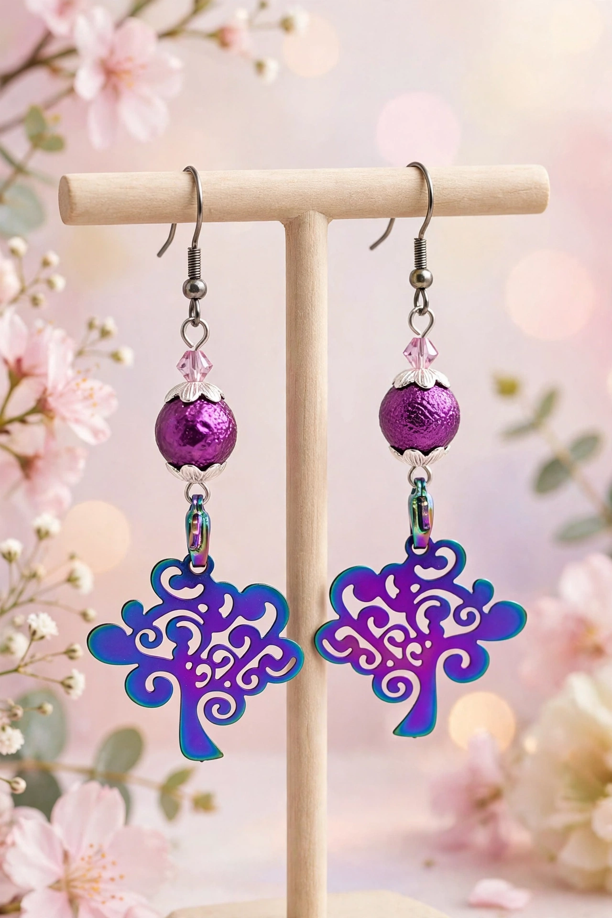 A pair of dangling earrings displayed on a wooden T-stand against a soft pink floral background. Each earring has a silver fishhook, a faceted purple bead, a small clear crystal, a metallic purple sphere, and a large tree-shaped drop with a purple-to-blue gradient finish.