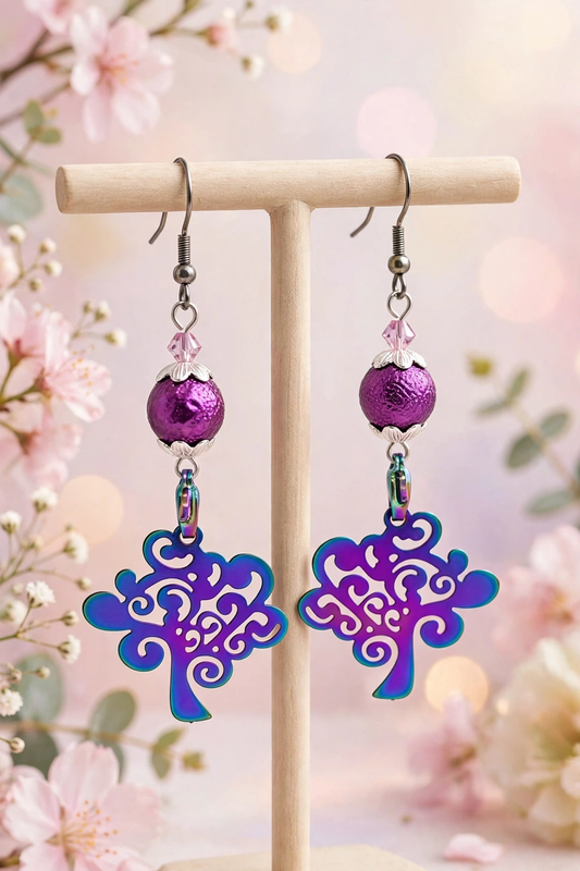 A pair of dangling earrings displayed on a wooden T-stand against a soft pink floral background. Each earring has a silver fishhook, a faceted purple bead, a small clear crystal, a metallic purple sphere, and a large tree-shaped drop with a purple-to-blue gradient finish.