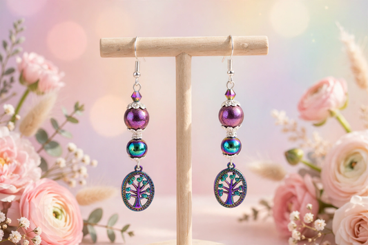 A pair of dangling earrings displayed on a wooden stand, featuring purple and iridescent blue-green beads with oval tree-of-life charms, set against a soft pink floral bokeh background.