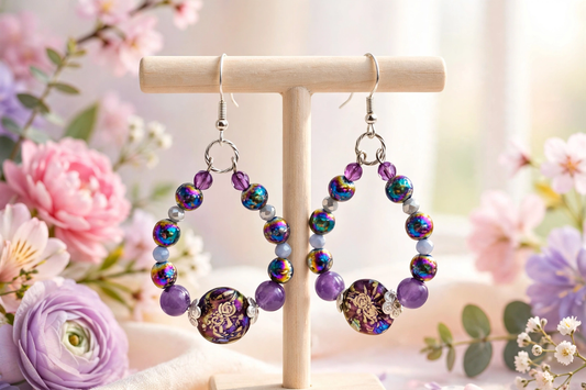 A pair of dangling beaded earrings displayed on a wooden stand, surrounded by soft pink and purple flowers. The earrings feature iridescent blue and purple beads, lavender beads, and a prominent round purple and gold patterned bead at the bottom.