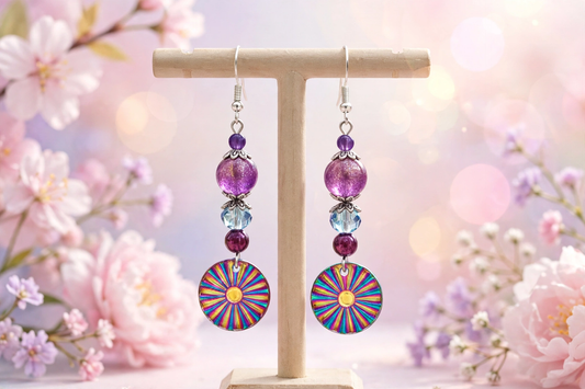 Colorful beaded dangle earrings displayed on a wooden T-stand, featuring purple and blue beads with circular sunburst-patterned charms, set against a soft pink floral bokeh background.
