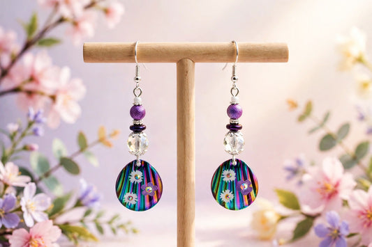 Handmade rainbow floral disc dangle earrings with purple beads on a wooden stand against blossoms