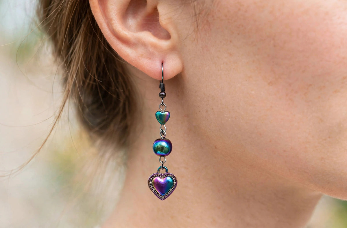 A close-up of a person's ear wearing a long dangling earring with three iridescent heart-shaped charms in purple, green, and blue tones.