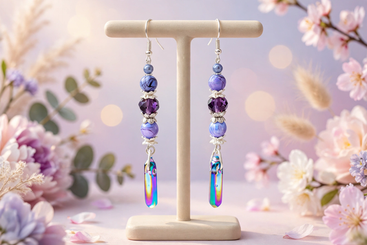 A pair of beaded dangle earrings displayed on a cream T-stand, set against a soft-focus floral background with pink blossoms and bokeh lights. The earrings feature purple and blue marbled beads, a purple faceted bead, silver-toned spacer beads, and a large iridescent crystal drop.