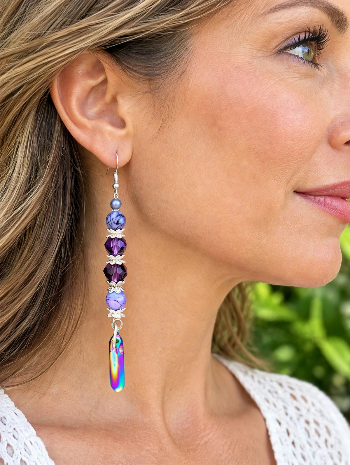 A close-up side profile of a woman wearing a long dangling earring. The earring features faceted purple beads, a small lavender bead, silver-toned metal spacers, and a large iridescent teardrop charm at the bottom.