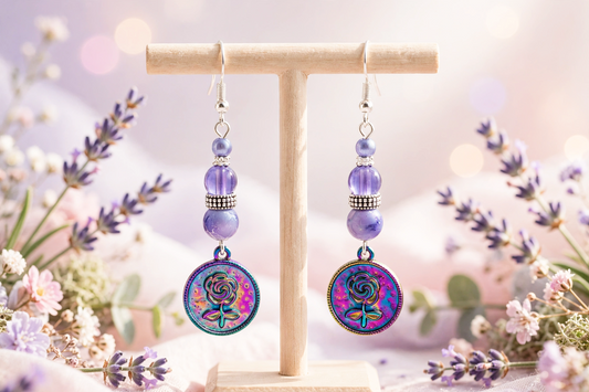 A pair of dangling earrings displayed on a small wooden T-stand, surrounded by soft lavender flowers and bokeh lights. The earrings feature silver hooks, purple beads, and round iridescent charms with a colorful floral pattern.