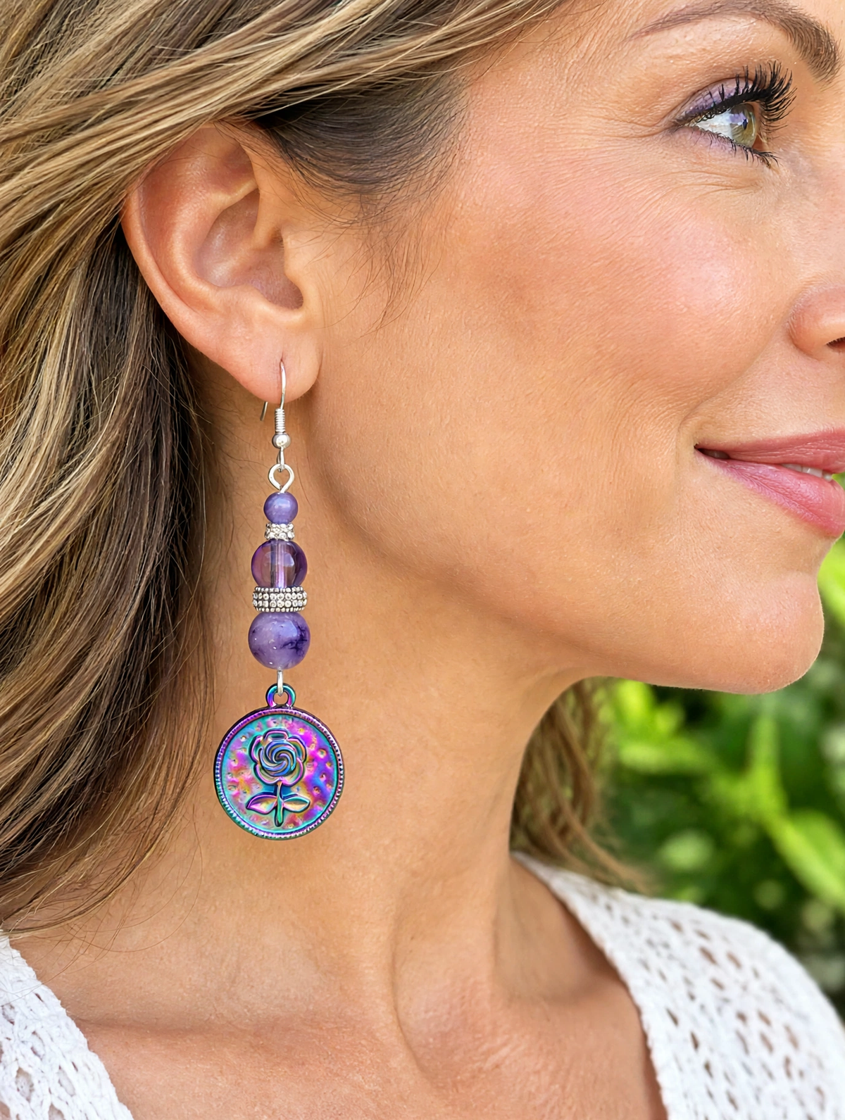 A close-up side profile of a woman wearing a dangling earring. The earring features purple beads, a silver-toned metal connector, and a round iridescent pendant with a colorful floral design in shades of blue, purple, and pink.