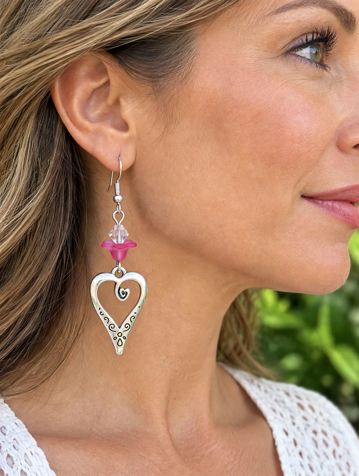 Woman wearing silver heart dangle earring with pink flower bead, outdoor lifestyle photo